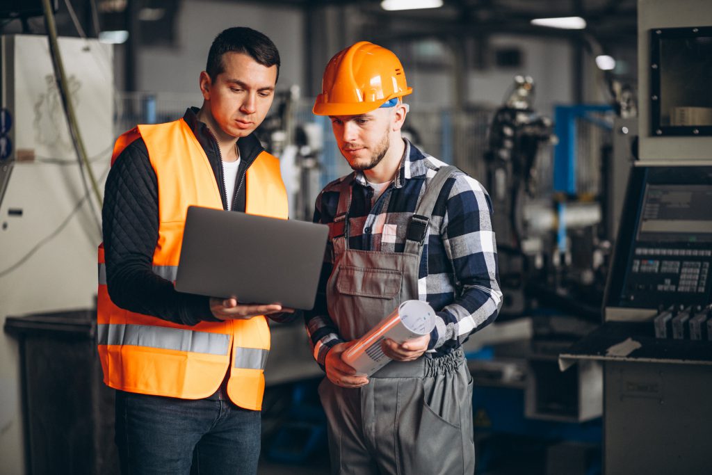 Dois profissionais em um ambiente industrial analisam informações em um notebook. À esquerda, uma pessoa veste colete de segurança laranja refletivo e segura o computador. À direita, outra pessoa usa capacete de segurança laranja e macacão de trabalho, segurando um documento enrolado. Ao fundo, há máquinas e equipamentos de uma fábrica, desfocados.
