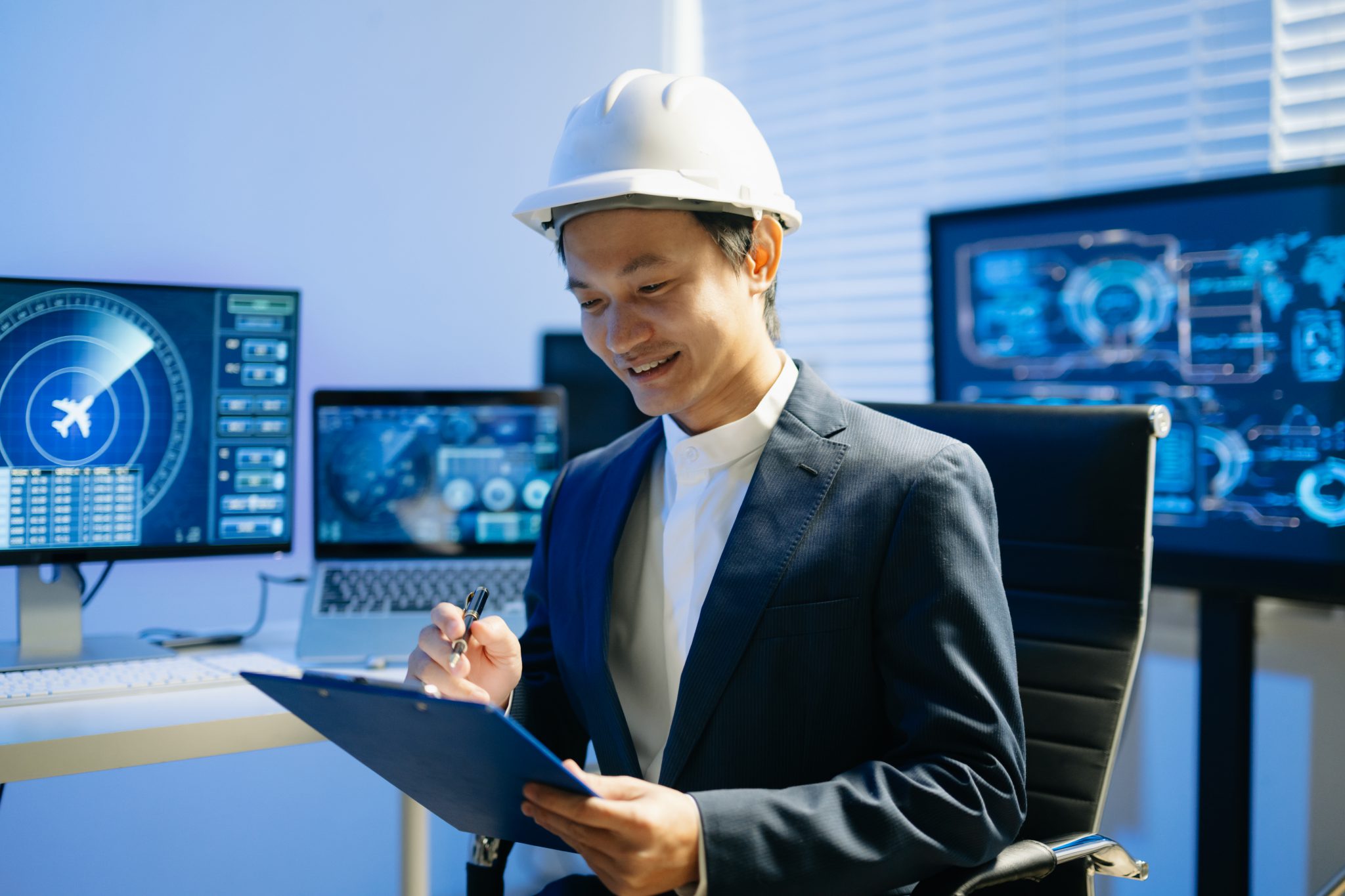 Engenheiro industrial usando capacete de segurança branco e segurando uma prancheta em uma sala de controle tecnológico, com monitores exibindo dados operacionais ao fundo.