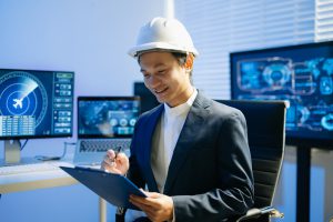 Engenheiro industrial usando capacete de segurança branco e segurando uma prancheta em uma sala de controle tecnológico, com monitores exibindo dados operacionais ao fundo.